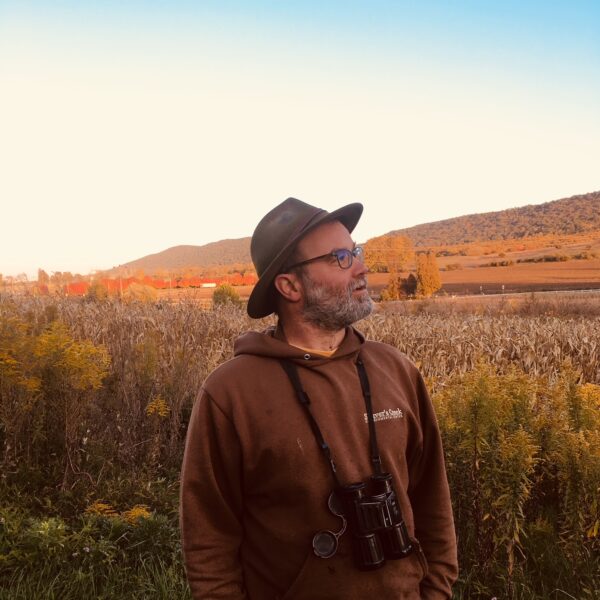 Jon, wearing a hat and binoculars stands in a field and gazes off into the distance.
