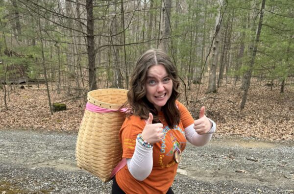 Brooke wears a woven backpack and give the double thumbs up as she walks along a wooded path.