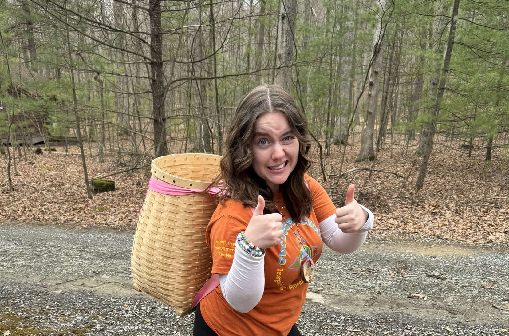 Brooke wears a woven backpack and give the double thumbs up as she walks along a wooded path.