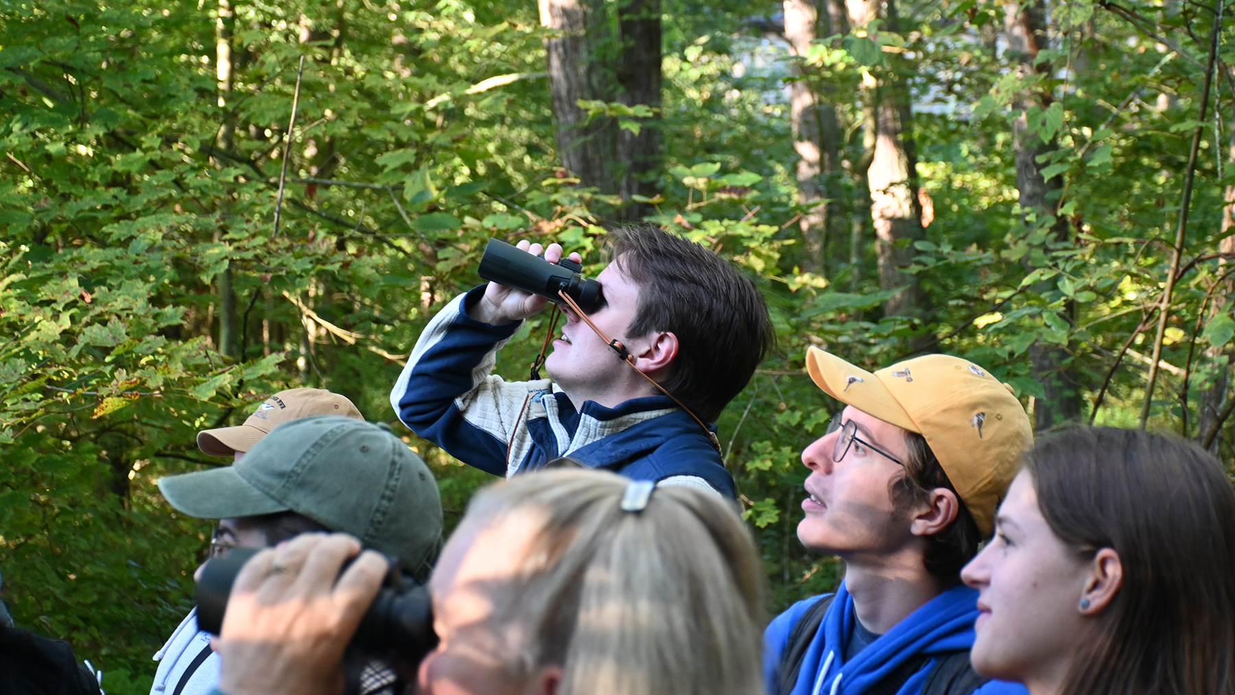 A group of people stand in the forest and watch for birds. One person is peering through binoculars.