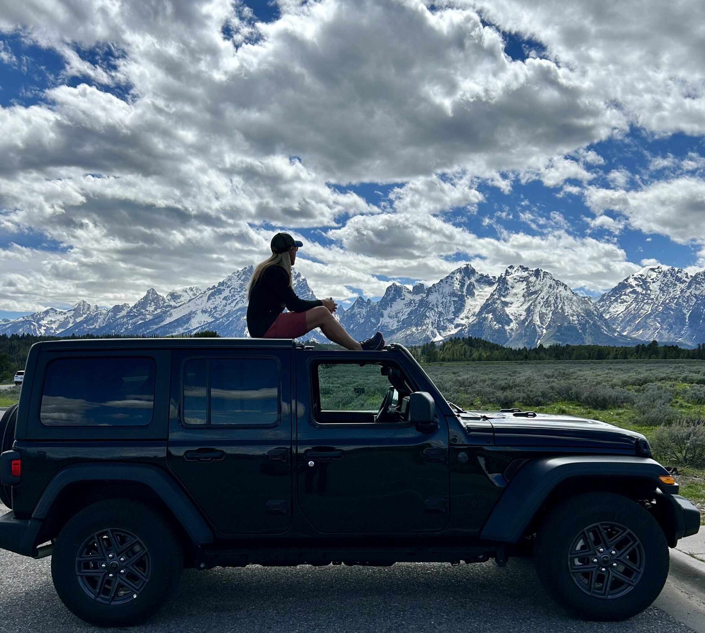 Nicole sitting on the roof of a jeep, silhouetted against the sky with a backdrop of snowy, rocky mountains.