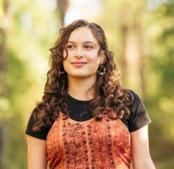 A young woman smiles wistfully in front of a blurred background of trees.