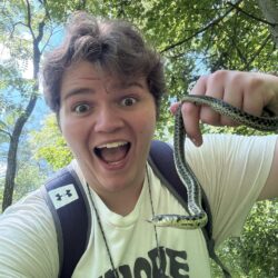A student wearing a backpack holds a small garter snake and smiles.